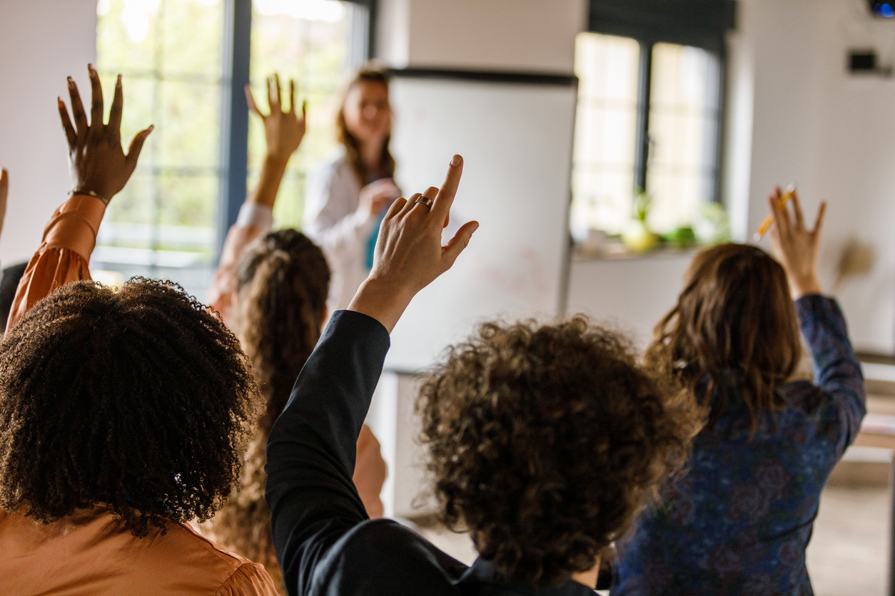 Diverse group of people raising hands in a classroom setting, seeking to answer question.